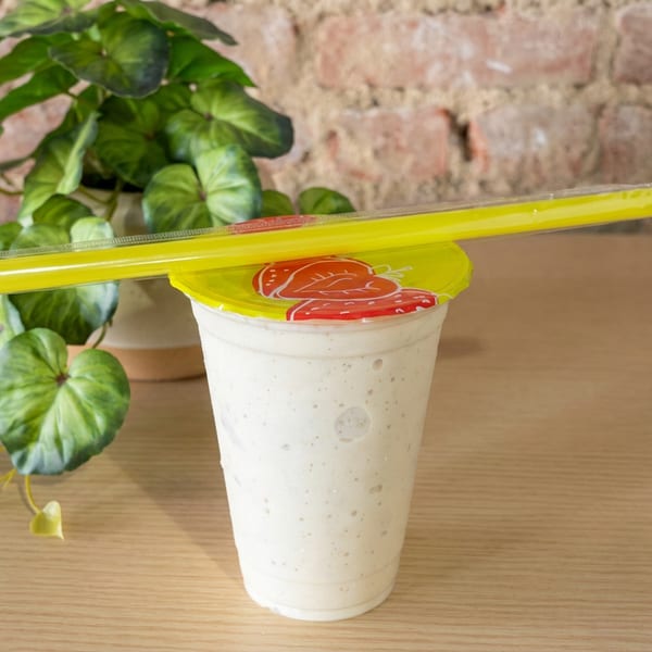 Creamy blended drink in a sealed plastic cup with a bright yellow straw, displayed on a wooden table beside a leafy plant.