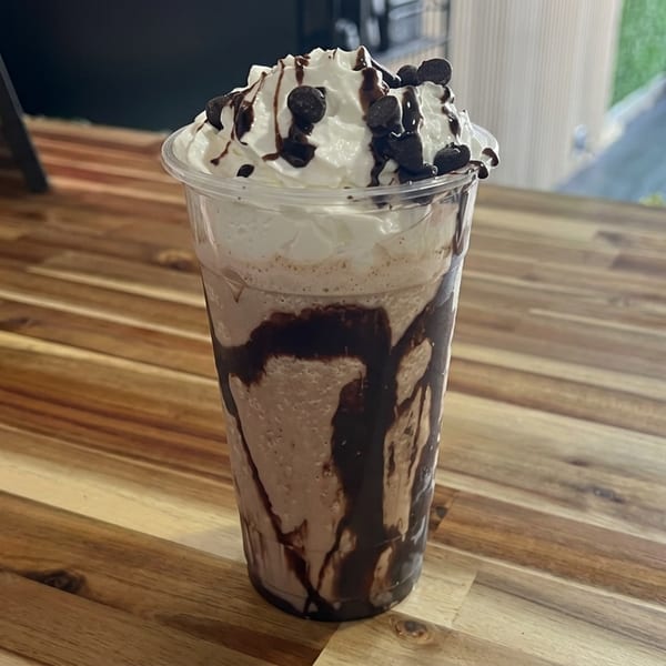 Chocolate milkshake topped with whipped cream, chocolate drizzle, and chocolate chips on a rustic wooden café table.