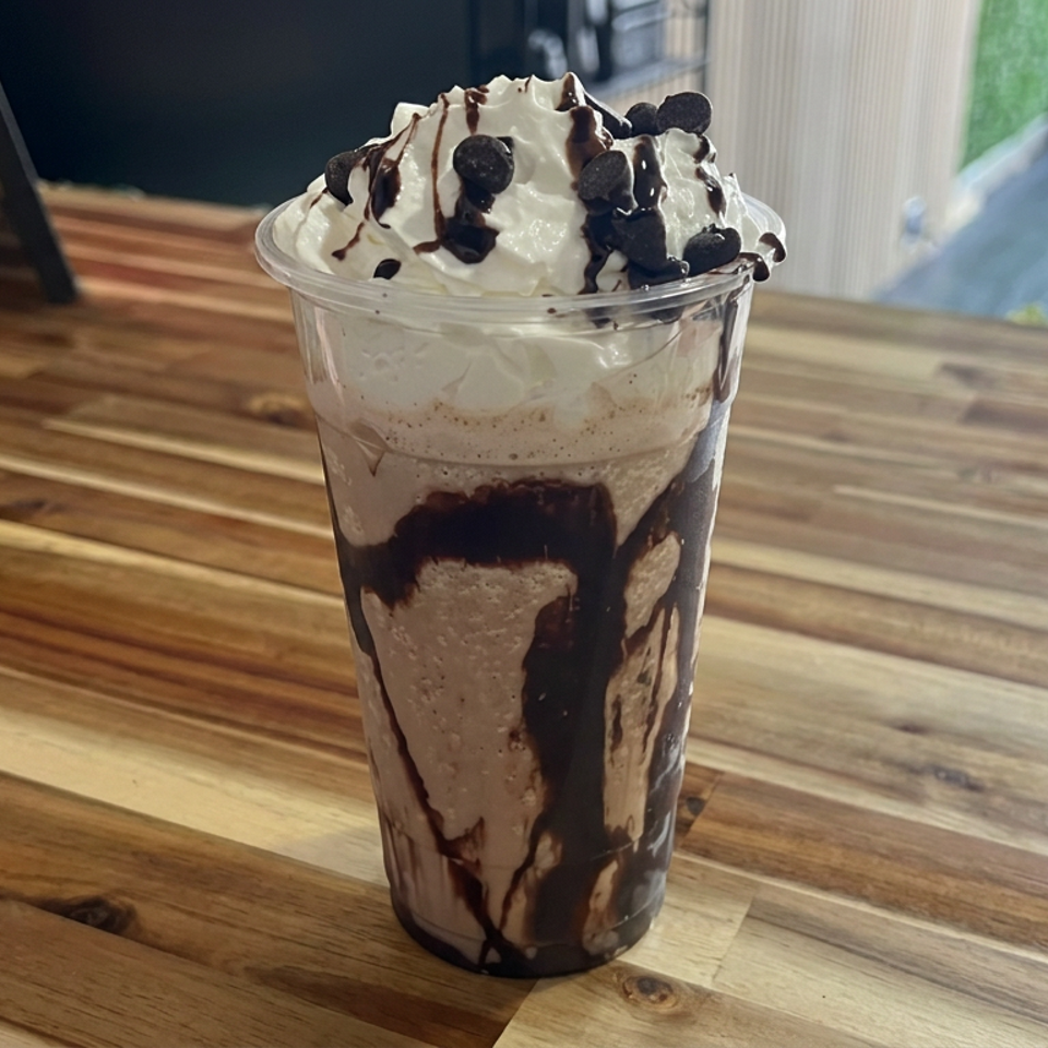 Chocolate milkshake topped with whipped cream, chocolate drizzle, and chocolate chips on a rustic wooden café table.
