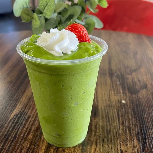 Green smoothie topped with whipped cream and a strawberry, served in a clear cup on a wooden restaurant table.