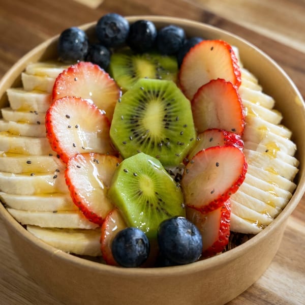 Fruit-topped smoothie bowl with sliced bananas, strawberries, kiwi, blueberries, chia seeds, and honey on a wooden table.