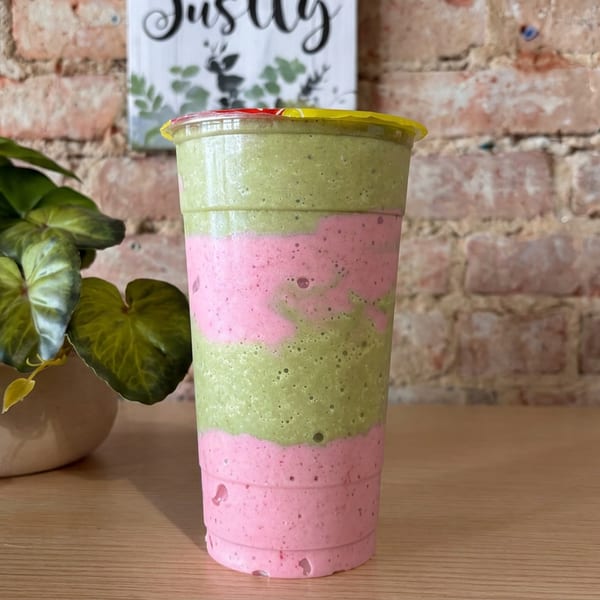 Layered green and pink smoothie in a clear cup on a wooden table beside a potted plant and brick wall.
