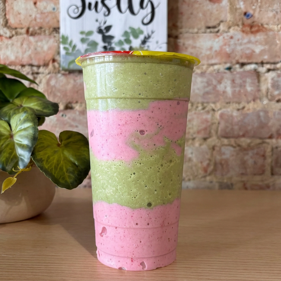 Layered pink and green smoothie in a clear cup sits on a wooden table beside a leafy potted plant and brick wall.
