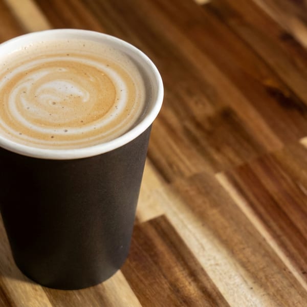 Foamy latte in a black takeaway cup sits on a sunlit rustic wooden table.